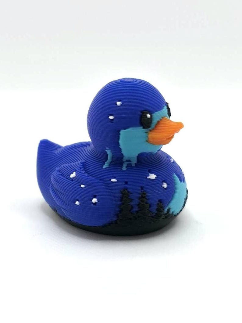 May include: A 3D printed rubber duck figure featuring a dark blue body with a light blue face and an orange beak. The design includes a black silhouette of a forest at the base, white star-like dots across the body, and visible horizontal layers.