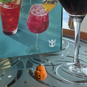 May include: A small orange pumpkin-shaped rubber duck figurine with a carved jack-o'-lantern face sits on a glass table next to a tall stemmed glass containing a dark liquid and a box featuring images of colorful cocktails and a white logo.