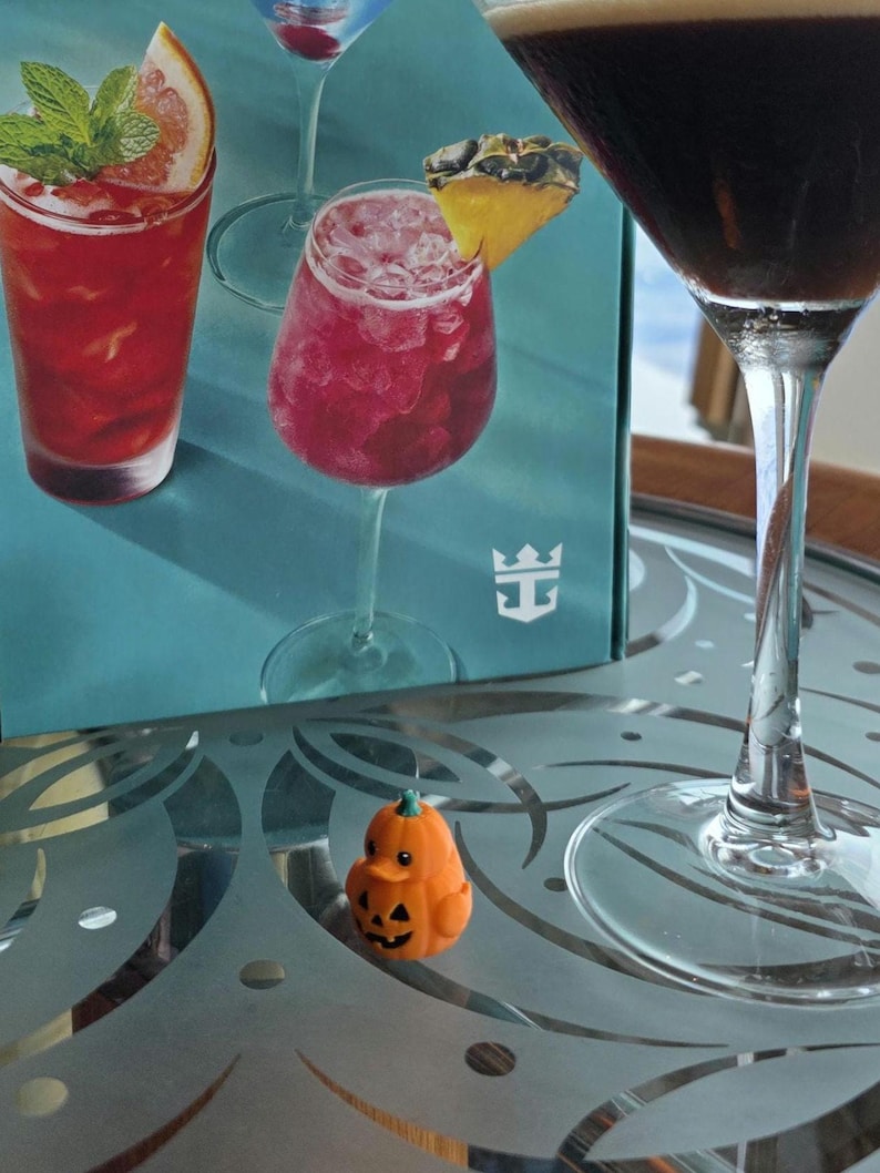 May include: A small orange pumpkin-shaped rubber duck figurine with a carved jack-o'-lantern face sits on a glass table next to a tall stemmed glass containing a dark liquid and a box featuring images of colorful cocktails and a white logo.