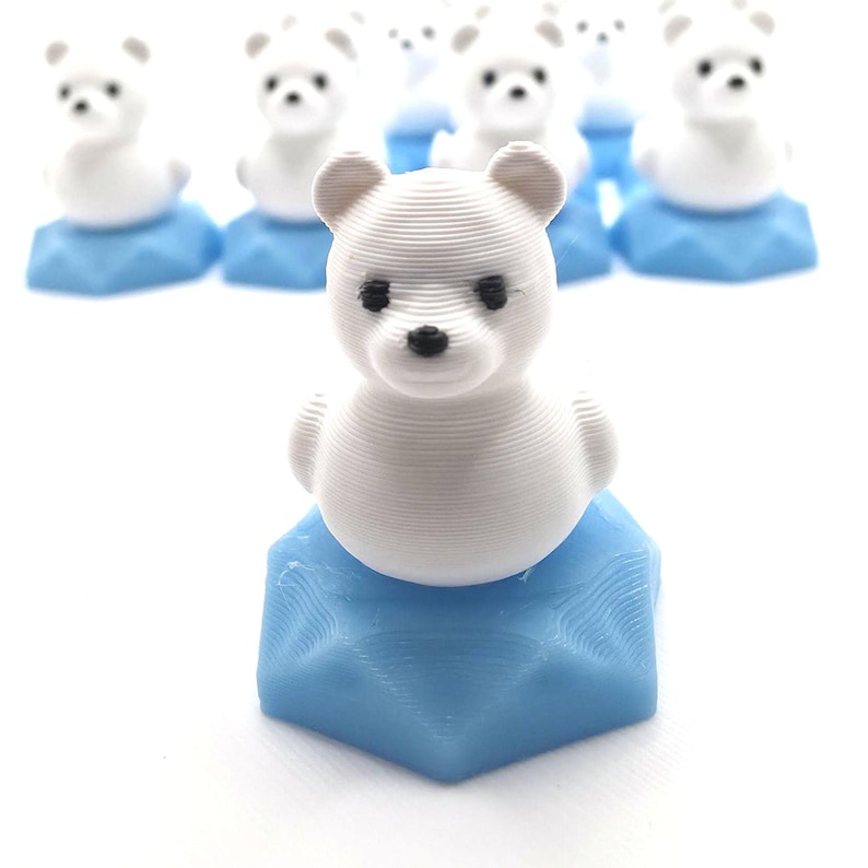 May include: A collection of small, white, 3D-printed polar bear figurines, each mounted on a light blue, hexagonal, textured base. The bears feature visible horizontal layer lines, black eyes, and a black nose, arranged in a group on a white surface.