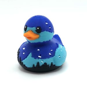 May include: A 3D printed rubber duck featuring a dark blue and light blue landscape design with a starry night sky, a black forest silhouette at the base, and a bright orange beak. The surface shows visible horizontal layer lines from the printing process.