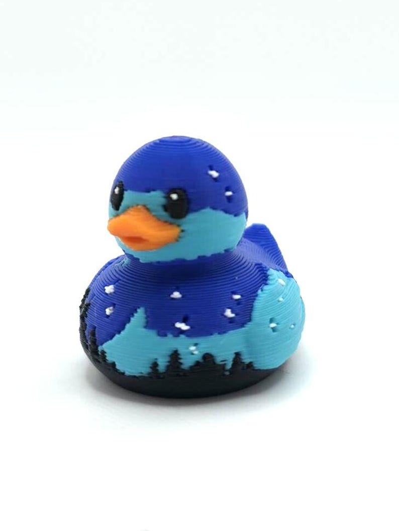 May include: A 3D printed rubber duck featuring a dark blue and light blue landscape design with a starry night sky, a black forest silhouette at the base, and a bright orange beak. The surface shows visible horizontal layer lines from the printing process.