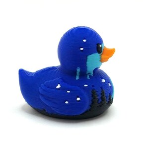 May include: A 3D printed rubber duck figure featuring a dark blue body with a light blue chest and an orange beak. The surface displays visible horizontal layer lines, with white speckles resembling snow and black silhouettes of pine trees.
