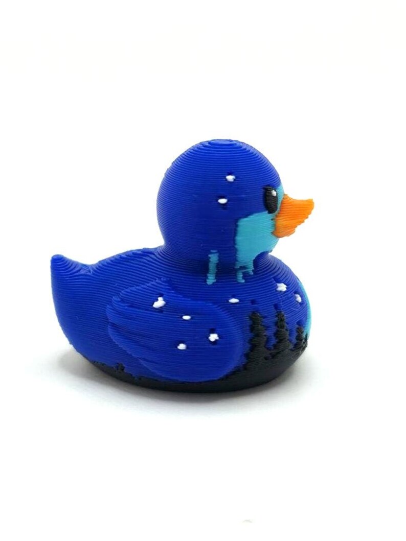 May include: A 3D printed rubber duck figure featuring a dark blue body with a light blue chest and an orange beak. The surface displays visible horizontal layer lines, with white speckles resembling snow and black silhouettes of pine trees.