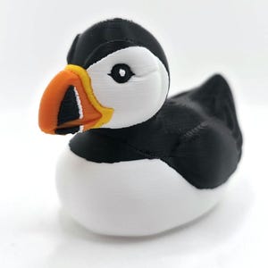 May include: A 3D printed puffin figurine featuring a black and white body with a distinctively colored beak. The beak is primarily orange with a yellow base and black and white markings. The bird has a white face, black head, and black wings.