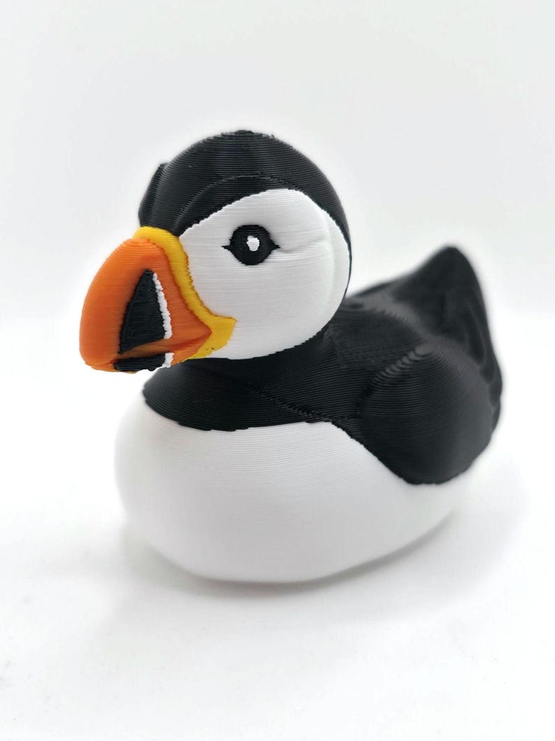 May include: A 3D printed puffin figurine featuring a black and white body with a distinctively colored beak. The beak is primarily orange with a yellow base and black and white markings. The bird has a white face, black head, and black wings.