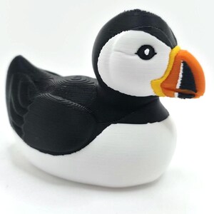 May include: A puffin figurine with a black head and back, white chest, and a large, colorful beak. The beak is orange, yellow, and black. The bird is sitting on a white surface with a blurred ocean background. 3d Printed in PLA Plastic.