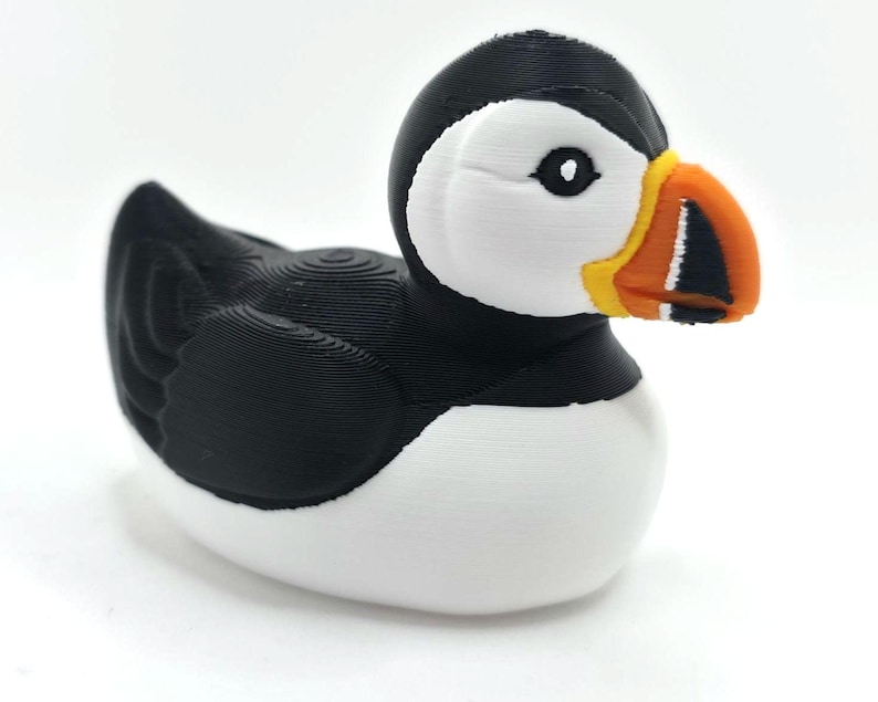 May include: A puffin figurine with a black head and back, white chest, and a large, colorful beak. The beak is orange, yellow, and black. The bird is sitting on a white surface with a blurred ocean background. 3d Printed in PLA Plastic.