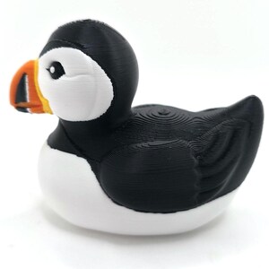 May include: A puffin figurine with a black head and back, white chest, and a large, colorful beak. The beak is orange, yellow, and black. The bird is sitting on a white surface with a blurred ocean background. 3d Printed in PLA Plastic.