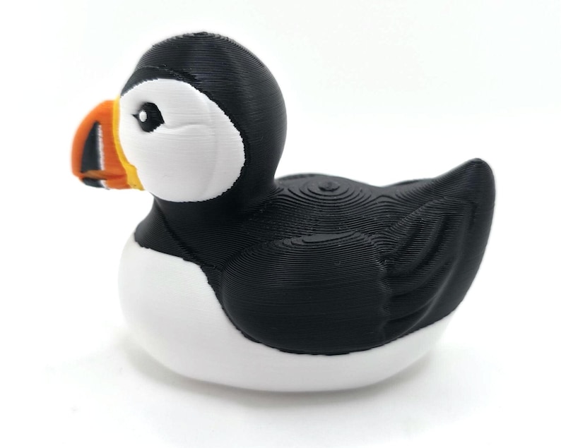 May include: A puffin figurine with a black head and back, white chest, and a large, colorful beak. The beak is orange, yellow, and black. The bird is sitting on a white surface with a blurred ocean background. 3d Printed in PLA Plastic.