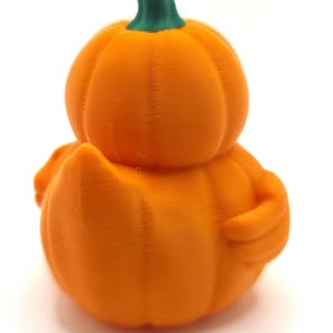 May include: A 3D printed orange pumpkin-shaped duck figurine with a green stem on top. The object features a rounded pumpkin body with visible horizontal layer lines, a small tail, and a wing-like protrusion on the side, all in a solid orange color.