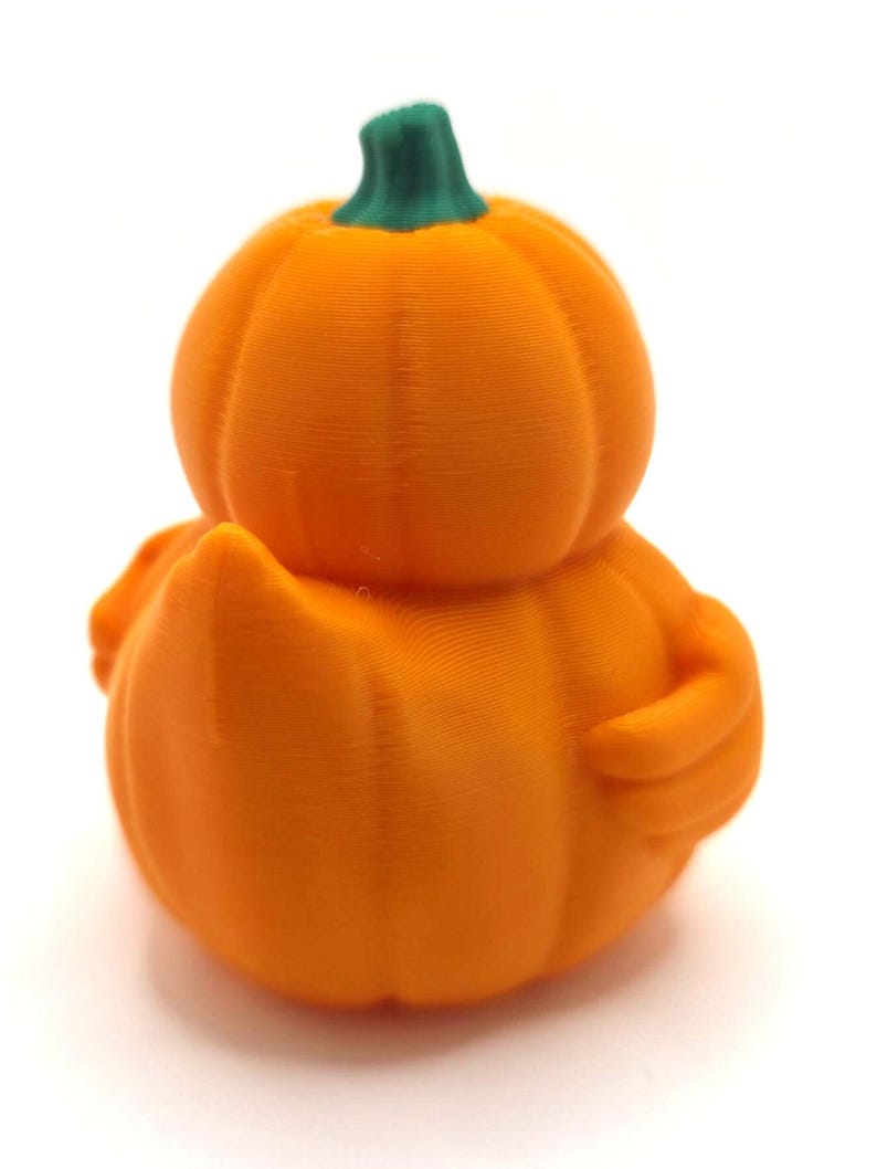 May include: A 3D printed orange pumpkin-shaped duck figurine with a green stem on top. The object features a rounded pumpkin body with visible horizontal layer lines, a small tail, and a wing-like protrusion on the side, all in a solid orange color.