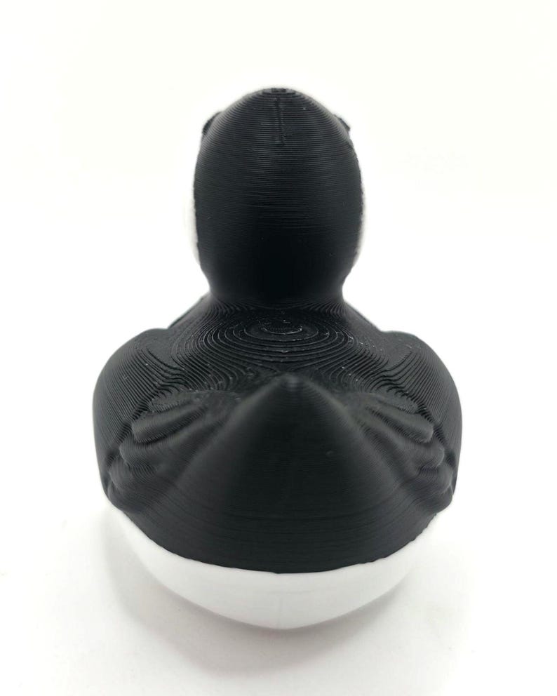 May include: A 3D printed duck figurine featuring a matte black head, neck, and back with visible horizontal layer lines. The lower portion of the duck is solid white, creating a distinct two-tone color scheme with a smooth, rounded base.