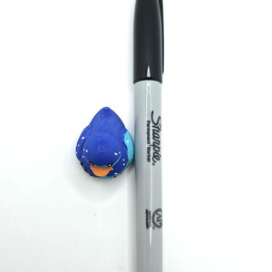 May include: A small blue rubber duck with white heart patterns and an orange beak positioned next to a grey and black Sharpie permanent marker for scale. The duck features a textured surface and dark eyes, while the marker displays the Sharpie logo.