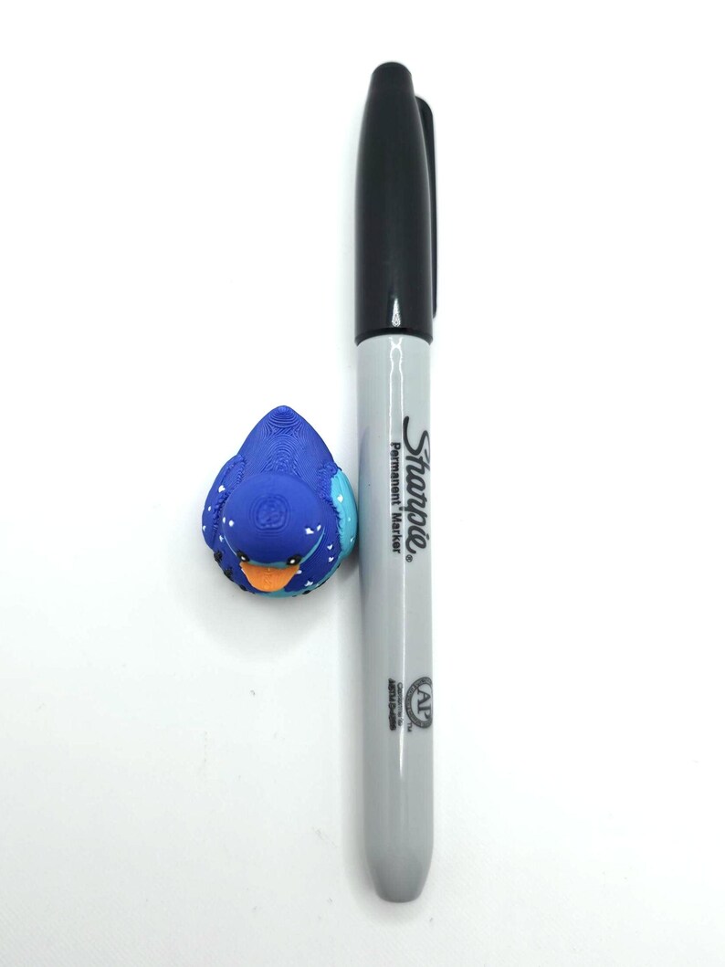 May include: A small blue rubber duck with white heart patterns and an orange beak positioned next to a grey and black Sharpie permanent marker for scale. The duck features a textured surface and dark eyes, while the marker displays the Sharpie logo.