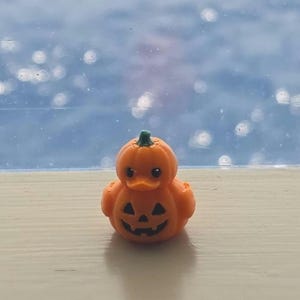 May include: A small orange rubber duck shaped like a pumpkin with a green stem on top. The duck features a carved jack-o'-lantern face with black triangular eyes and a jagged mouth. The surface has a visible 3D printed texture throughout the body.