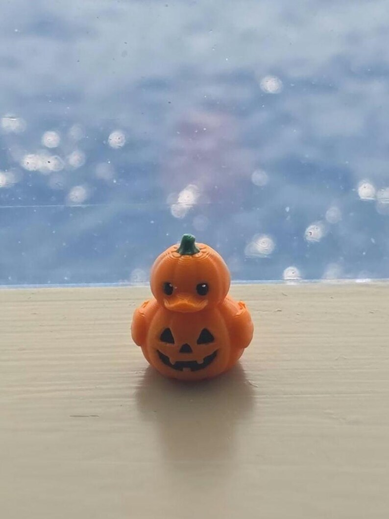 May include: A small orange rubber duck shaped like a pumpkin with a green stem on top. The duck features a carved jack-o'-lantern face with black triangular eyes and a jagged mouth. The surface has a visible 3D printed texture throughout the body.