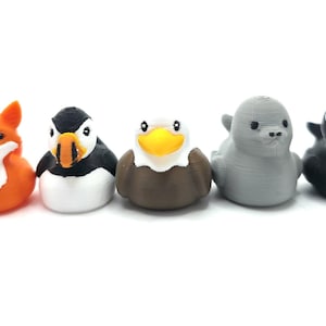 Puede incluir: Un conjunto de seis peque&ntilde;as figuras con forma de animal dispuestas en fila. La colecci&oacute;n incluye un lobo gris, un zorro naranja, un frailecillo blanco y negro, un &aacute;guila marr&oacute;n y blanca con pico amarillo, una foca gris s&oacute;lida y una orca blanca y negra.