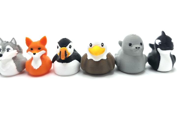 Puede incluir: Un conjunto de seis peque&ntilde;as figuras con forma de animal dispuestas en fila. La colecci&oacute;n incluye un lobo gris, un zorro naranja, un frailecillo blanco y negro, un &aacute;guila marr&oacute;n y blanca con pico amarillo, una foca gris s&oacute;lida y una orca blanca y negra.