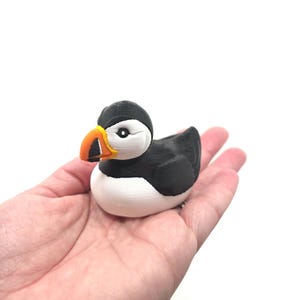 May include: A small, three-dimensional printed figurine of a puffin bird. The figure features a black head, back, and wings, with a white chest and face. It has a distinct, bright orange and yellow beak with black detailing around the base.