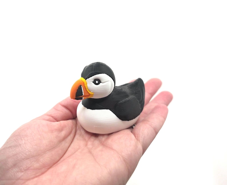 May include: A small, three-dimensional printed figurine of a puffin bird. The figure features a black head, back, and wings, with a white chest and face. It has a distinct, bright orange and yellow beak with black detailing around the base.