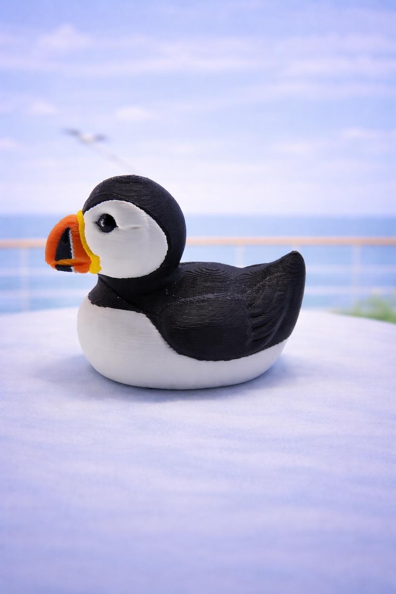 May include: A puffin figurine with a black head and back, white chest, and a large, colorful beak. The beak is orange, yellow, and black. The bird is sitting on a white surface with a blurred ocean background. 3d Printed in PLA Plastic.