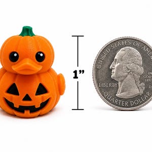 May include: An orange pumpkin-shaped rubber duck with a green stem on top, black eyes, and a carved jack-o'-lantern face on the body. A vertical line with the text 1 inch indicates the height next to a silver United States quarter dollar coin.