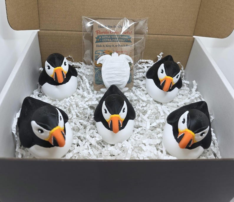 May include: A cardboard box containing four black and white puffin figurines with orange beaks, nestled in white paper shred. A small clear plastic bag holds a white turtle figurine and a card with the text Florida Cruise Ducks A Little Something Extra For You.