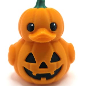 May include: A small orange rubber duck shaped like a pumpkin with a green stem on top. The duck features a carved jack-o'-lantern face with black triangular eyes and a jagged mouth. The surface has a visible 3D printed texture throughout.