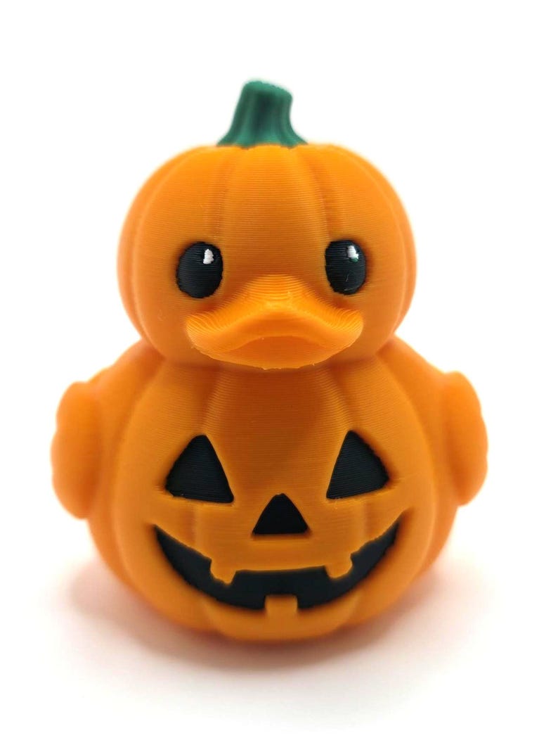 May include: A small orange rubber duck shaped like a pumpkin with a green stem on top. The duck features a carved jack-o'-lantern face with black triangular eyes and a jagged mouth. The surface has a visible 3D printed texture throughout.