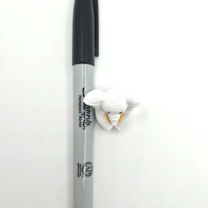 May include: A gray Sharpie permanent marker positioned vertically next to a small, white, textured elephant figurine. The marker features a black cap and black text on the barrel, while the elephant has visible ear details and a yellow accent.