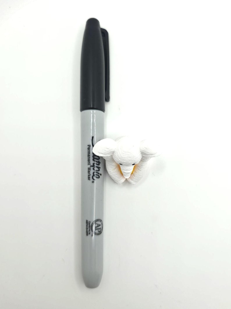 May include: A gray Sharpie permanent marker positioned vertically next to a small, white, textured elephant figurine. The marker features a black cap and black text on the barrel, while the elephant has visible ear details and a yellow accent.