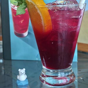 May include: A tall glass filled with a deep red beverage, ice cubes, and a white straw. The drink is garnished with a bright orange slice and fresh green mint leaves. Beside the glass sits a small, white, cartoon-style bear figurine on a blue base.