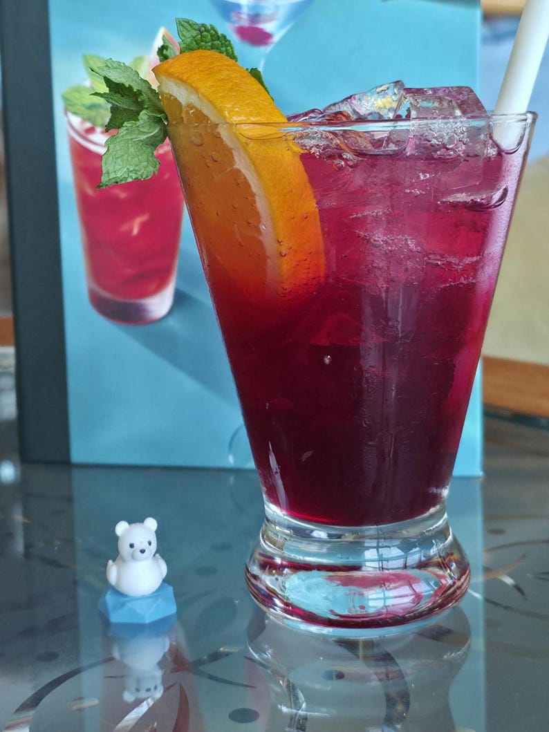 May include: A tall glass filled with a deep red beverage, ice cubes, and a white straw. The drink is garnished with a bright orange slice and fresh green mint leaves. Beside the glass sits a small, white, cartoon-style bear figurine on a blue base.