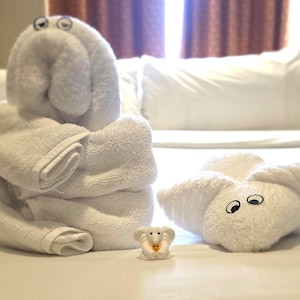 May include: Two white towel sculptures shaped like animals with attached googly eyes, one standing upright and one lying flat, positioned next to a small white elephant figurine holding a tiny yellow object in its trunk.
