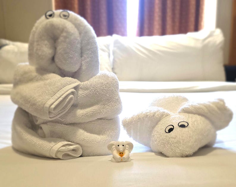 May include: Two white towel sculptures shaped like animals with attached googly eyes, one standing upright and one lying flat, positioned next to a small white elephant figurine holding a tiny yellow object in its trunk.