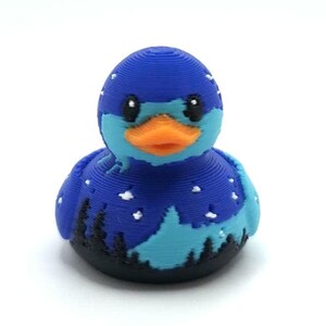 May include: A 3D printed rubber duck featuring a dark blue night sky pattern with white stars, a light blue mountain silhouette, and a black forest landscape at the base. The duck has a bright orange beak and black eyes, with visible horizontal layer lines.