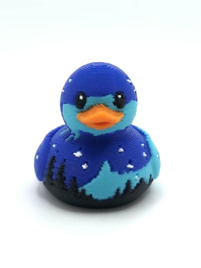 May include: A 3D printed rubber duck featuring a dark blue night sky pattern with white stars, a light blue mountain silhouette, and a black forest landscape at the base. The duck has a bright orange beak and black eyes, with visible horizontal layer lines.