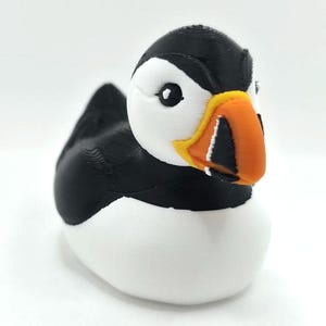 May include: A 3D printed puffin figurine featuring a black back, wings, and head cap, with a white face, chest, and belly. The bird has a distinct orange, yellow, and black beak with visible horizontal ridges and a small black eye on the white face.