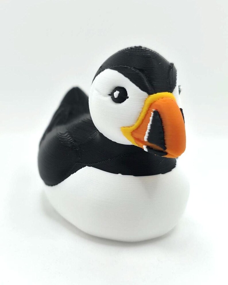 May include: A 3D printed puffin figurine featuring a black back, wings, and head cap, with a white face, chest, and belly. The bird has a distinct orange, yellow, and black beak with visible horizontal ridges and a small black eye on the white face.