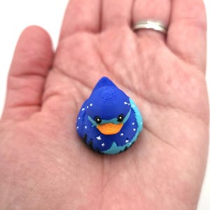 May include: A small, 3D-printed duck figurine featuring a deep blue body decorated with white star patterns and a silhouette of black trees along the base. The duck has a bright orange beak and black eyes, showcasing a layered texture from the printing process.