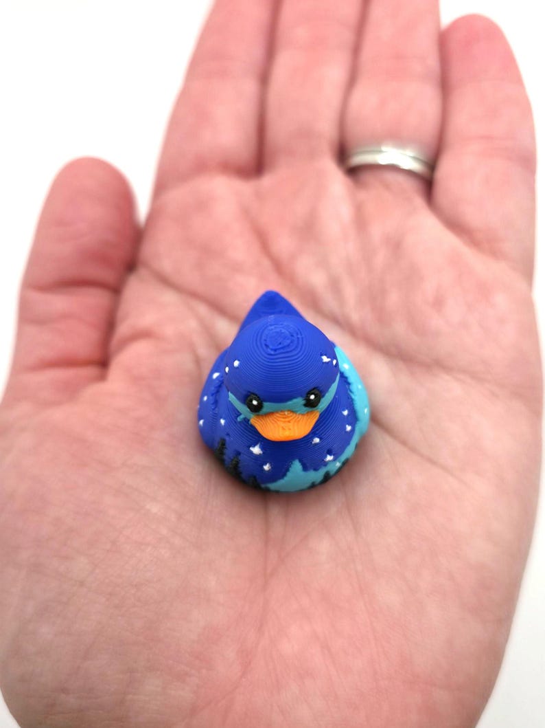 May include: A small, 3D-printed duck figurine featuring a deep blue body decorated with white star patterns and a silhouette of black trees along the base. The duck has a bright orange beak and black eyes, showcasing a layered texture from the printing process.