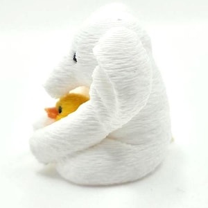 May include: A white 3D printed elephant figurine with a textured, layered surface. The elephant is depicted in a seated position, holding a small, bright yellow duckling with an orange beak tucked against its chest. The elephant has a black eye.