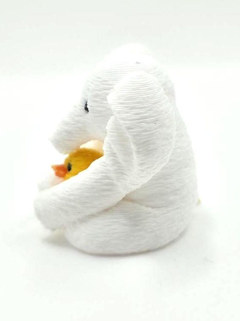 May include: A white 3D printed elephant figurine with a textured, layered surface. The elephant is depicted in a seated position, holding a small, bright yellow duckling with an orange beak tucked against its chest. The elephant has a black eye.