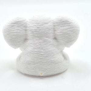 May include: A white 3D printed figurine shaped like an elephant head with large rounded ears. The object features a distinct layered texture from the printing process and a small yellow dot at the base of the front center of the structure.