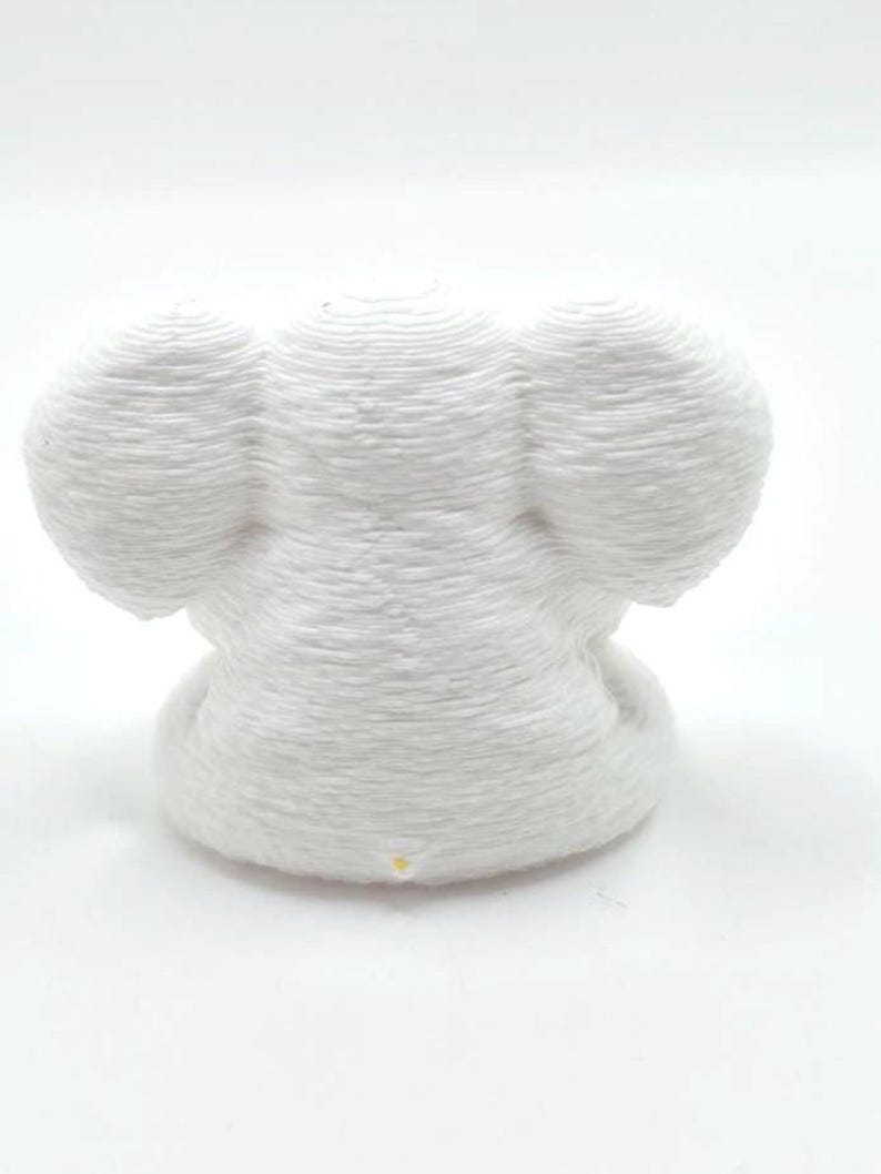 May include: A white 3D printed figurine shaped like an elephant head with large rounded ears. The object features a distinct layered texture from the printing process and a small yellow dot at the base of the front center of the structure.