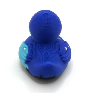 May include: A 3D printed duck figurine featuring a layered texture throughout its surface. The duck is primarily deep blue with light blue accents on the wings and small white details on the wing tips, viewed from the back against a white background.