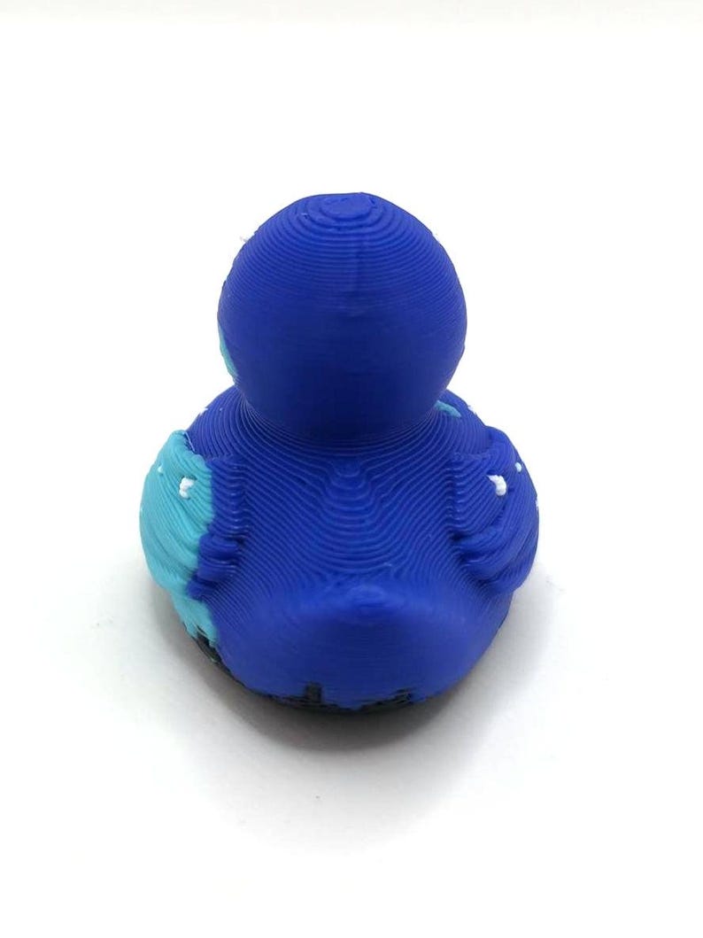 May include: A 3D printed duck figurine featuring a layered texture throughout its surface. The duck is primarily deep blue with light blue accents on the wings and small white details on the wing tips, viewed from the back against a white background.