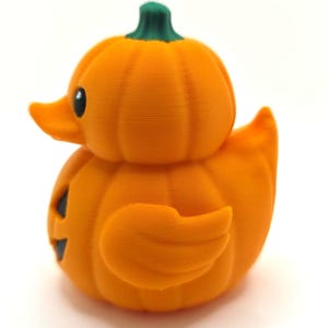 May include: A small orange duck-shaped figurine with a pumpkin-like ribbed texture, a green stem on top, and a black eye. The duck has a flat orange beak and a carved black jack-o'-lantern face on its side, featuring a triangular nose and mouth.