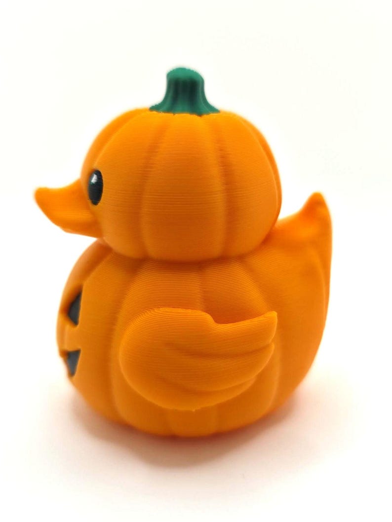 May include: A small orange duck-shaped figurine with a pumpkin-like ribbed texture, a green stem on top, and a black eye. The duck has a flat orange beak and a carved black jack-o'-lantern face on its side, featuring a triangular nose and mouth.